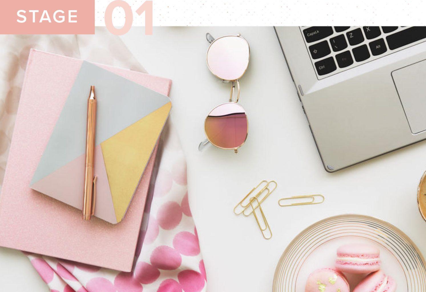 a desk with a pink notebook, pink sunglasses, macarons, and a laptop