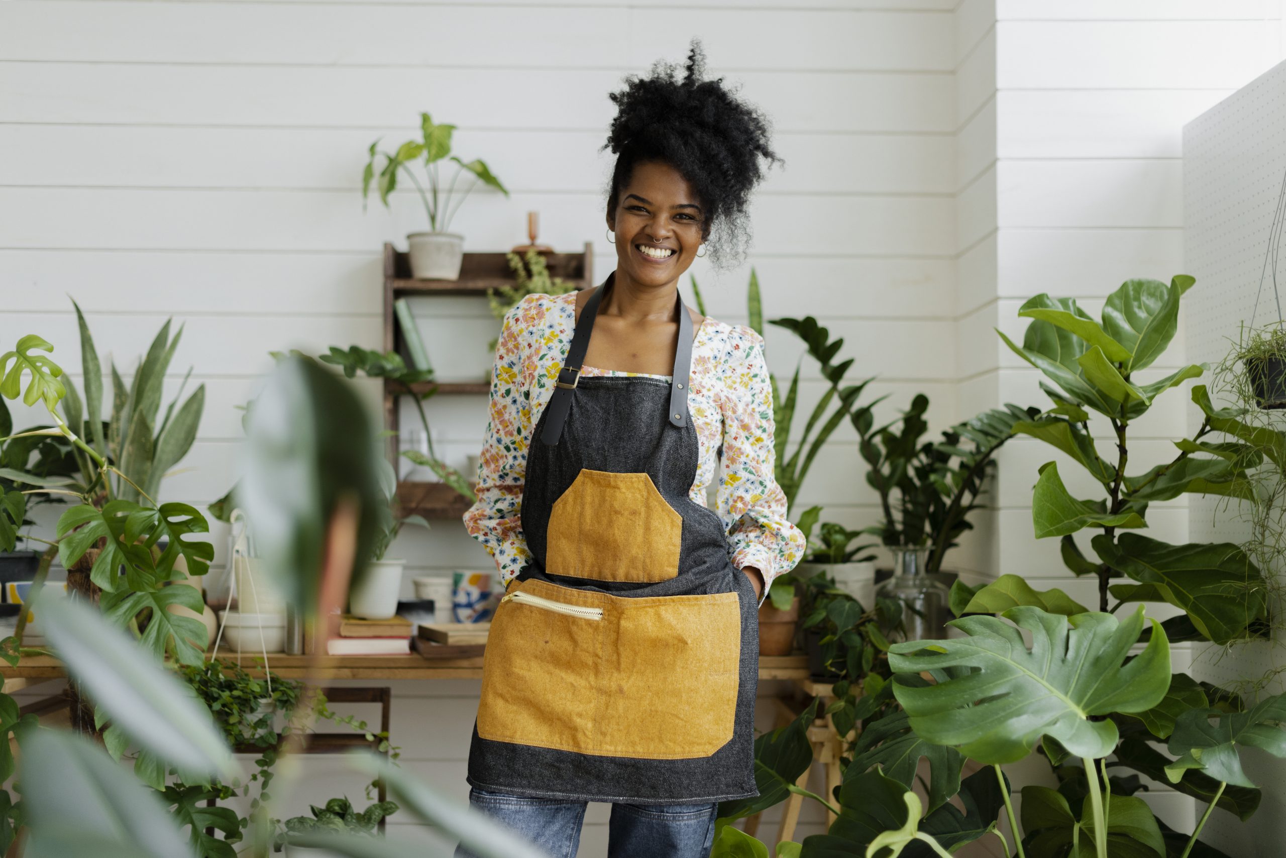 A woman smiling at the camera and standing in her indoor garden wearing gardening equipments